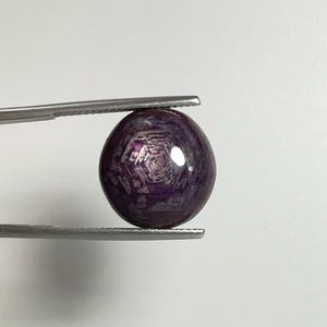 May include: A spherical, polished gemstone held by tweezers. The gem is a deep purple colour with a star-like pattern visible on its surface. The gem is smooth and reflective, with a slight sheen. The background is plain white.