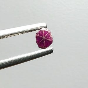 May include: A small, faceted, deep red gemstone is held by silver tweezers. The gemstone has a star-like pattern on its surface, set against a plain, light background.
