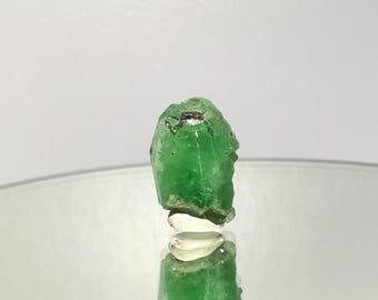 May include: A vibrant green gemstone with a rough, textured surface. The gemstone is mounted on a clear, reflective base, creating a striking contrast. The stone's color is a rich, saturated green, and it appears to be a natural, uncut crystal.