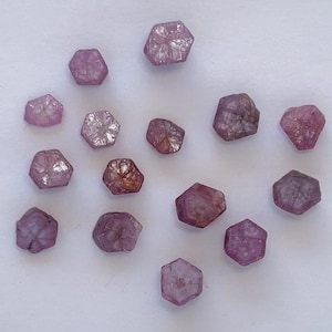 May include: A selection of pink and purple hexagonal gemstones. The raw crystals have a natural, unpolished appearance, with variations in colour and clarity. The gemstones are scattered on a white surface.