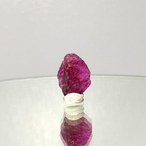 Peut inclure: Une pierre précieuse de rubis brute, non taillée, d'un magenta vif. La gemme est montée sur une bague en argent, se reflétant sur une surface miroir. La texture brute et la couleur du rubis sont mises en évidence.