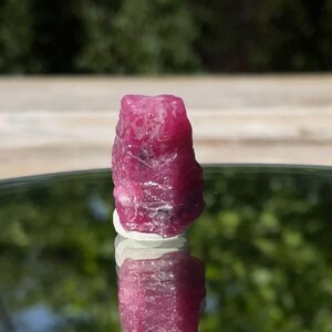 May include: A raw, unpolished ruby crystal with a deep magenta-red hue and a textured, multifaceted surface. The stone features natural crystalline formations and irregular edges, displaying a vibrant, opaque color throughout its vertical structure.