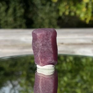 Peut inclure: Un cristal de rubis brut et non poli avec une teinte magenta-rose profonde et une structure colonnaire hexagonale. La pierre présente une surface cristalline texturée avec des facettes naturelles et repose sur une petite base circulaire blanche.