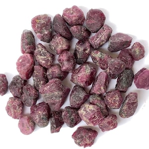 May include: A collection of rough, pink ruby gemstones. The rubies are various sizes and shapes, with some having a darker, more purplish hue.