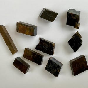 May include: A collection of ten black and brown crystals of various shapes and sizes. The crystals are arranged on a white surface.