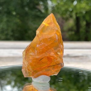 May include: A translucent, orange-yellow crystal formation. The rough-hewn stone is displayed on a reflective surface, with a blurred green background. The crystal's irregular shape and color variations suggest a natural, unpolished state.
