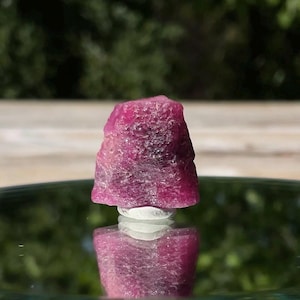 May include: A raw, unpolished ruby gemstone with a deep magenta and pink hue. The stone features a rough, crystalline texture with natural facets and irregular edges, displaying a vibrant, saturated colour throughout its solid, opaque structure.