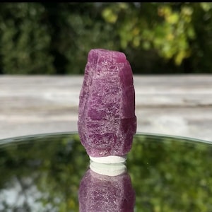 May include: A raw, deep magenta-coloured crystal specimen with a hexagonal, columnar structure. The surface of the stone displays a rough, textured appearance with natural facets and internal fractures, reflecting light across its translucent body.