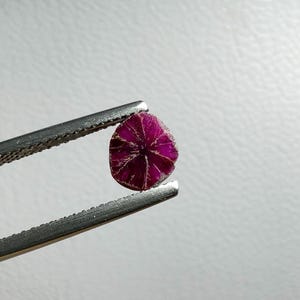 May include: A small, round, deep magenta gemstone is held by silver tweezers. The gemstone features a star-like pattern of lighter lines radiating from the centre. The background is a plain, off-white surface.
