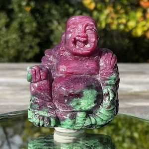 May include: A hand-carved figurine of a laughing Buddha made from a combination of deep magenta ruby and vibrant green zoisite. The stone features a polished texture with visible natural color variations throughout the seated figure.