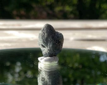 May include: A dark gray, rough-textured stone, possibly a mineral specimen, is displayed on a small white base. The stone is reflected in a shiny, dark surface, creating a mirrored effect. The background is blurred.