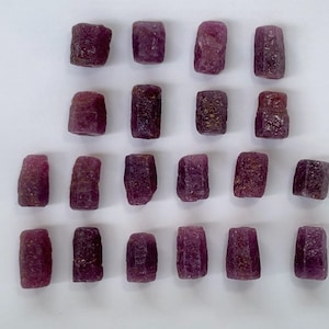 May include: A collection of raw ruby gemstones. The stones are a deep red-purple colour, with a rough, textured surface. The gemstones are rectangular and cylindrical in shape, and are arranged on a white surface.