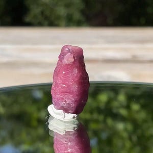 May include: A raw, unpolished ruby crystal with a deep magenta-pink hue and a textured, irregular surface. The stone stands upright on a small white base, displaying a natural, crystalline structure with visible facets and opaque sections.