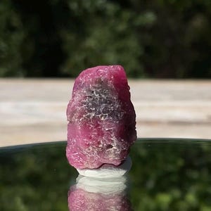 May include: A raw, unpolished ruby crystal with a deep magenta and pink hue. The stone features a rough, textured surface with natural crystalline formations and dark inclusions visible across its irregular, blocky, and vertical structure.