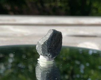 May include: A dark gray, rough-textured mineral specimen, possibly a crystal, is displayed. The stone is set on a reflective surface, with a blurred background of green and white. The mineral's shape is irregular, with visible facets.
