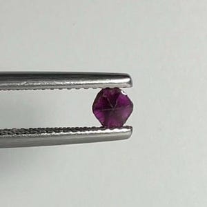 May include: A faceted, deep purple gemstone is held by a pair of silver tweezers against a white background. The gemstone has multiple facets, reflecting light and showcasing its rich color. The tweezers provide a secure grip on the gem.