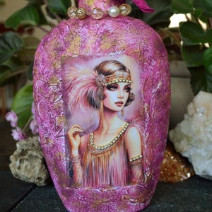 May include: A decorative, pink bottle with a faceted, magenta stopper. The bottle is adorned with a portrait of a woman in a flapper-style dress. The bottle's neck is wrapped with a magenta ribbon and embellished with beads.