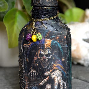 May include: A decorative glass bottle with a textured black finish featuring a graphic of a jester with a pointed hat and reaching hand. The bottle is adorned with a gold chain holding three small purple, yellow, and green skull charms.