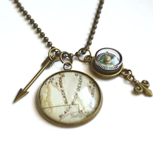 May include: A bronze necklace with a compass, an arrow charm, and a round pendant with a map of Black Rock Desert, Nevada. The compass has a green needle and a clear face.