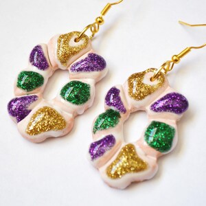 May include: A pair of handmade earrings featuring a pink, donut-shaped design with gold, green, and purple glitter accents. The earrings have gold-colored fish hook ear wires.