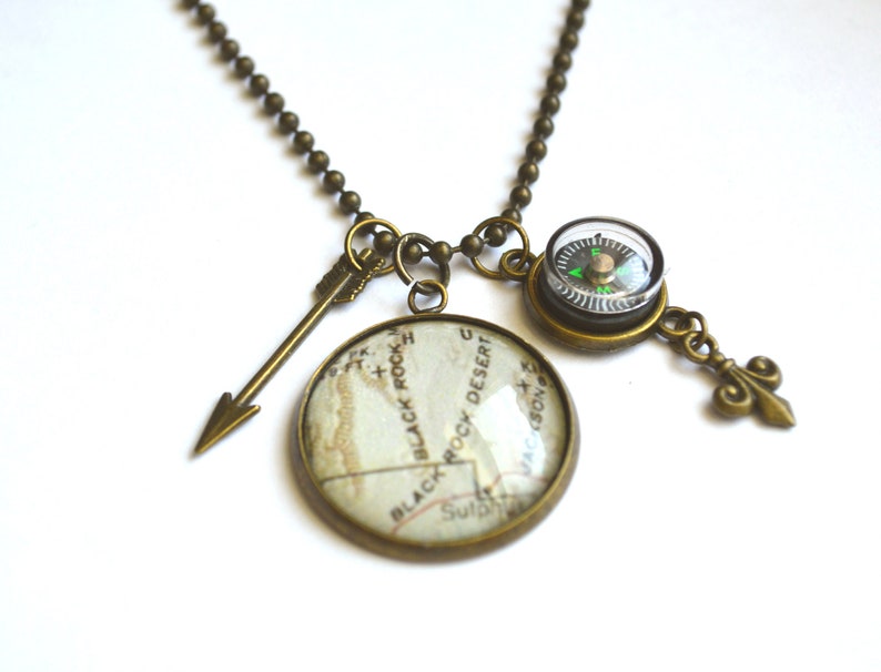 May include: A bronze necklace with a compass, an arrow charm, and a round pendant with a map of Black Rock Desert, Nevada. The compass has a green needle and a clear face.