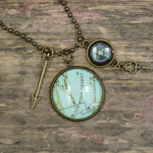 May include: A necklace with a compass, a key charm, and a round glass pendant with a map of Black Rock Desert, Utah. The necklace is made of a bronze chain.