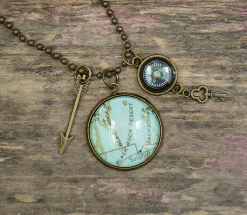 May include: A necklace with a compass, a key charm, and a round glass pendant with a map of Black Rock Desert, Utah. The necklace is made of a bronze chain.