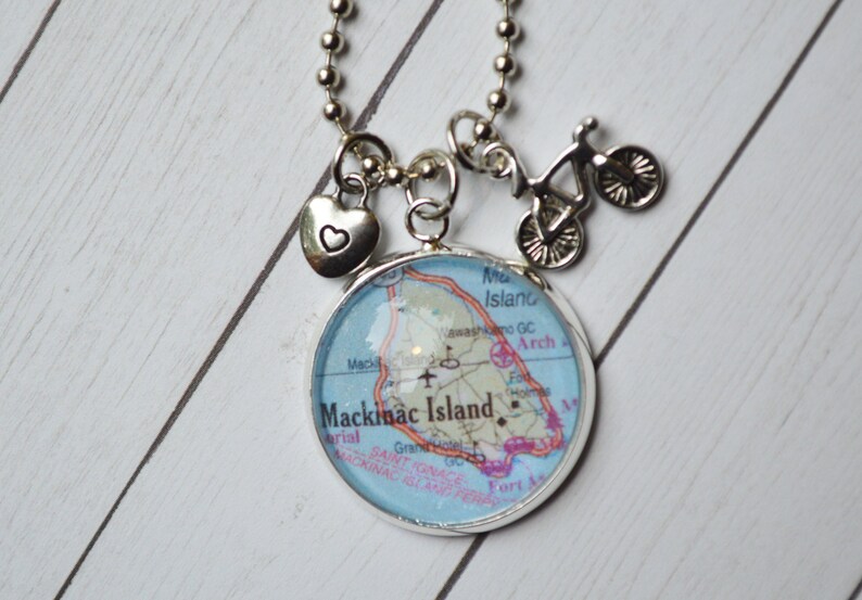 Op de afbeelding: Een zilveren ketting met een bedel met een kaart van Mackinac Island, Michigan. De ketting heeft ook een klein hartbedel en een fietsbedel.