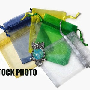 Op de afbeelding: Assortiment van kleurrijke organza-geschenkzakjes in geel, groen, blauw en zilver. De zakjes zijn gemaakt van transparante stof en hebben een trekkoord. De zakjes zijn in een stapel gerangschikt met een zilveren uilbedel bovenop.