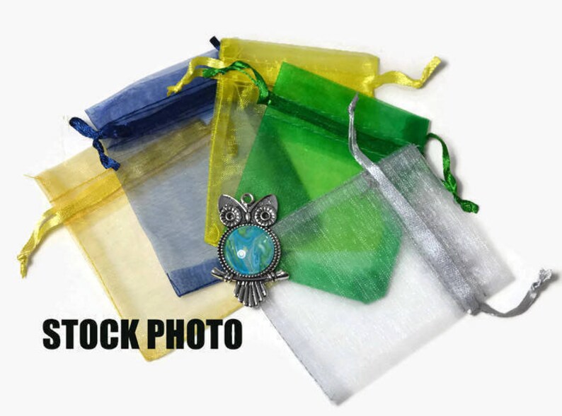 Op de afbeelding: Assortiment van kleurrijke organza-geschenkzakjes in geel, groen, blauw en zilver. De zakjes zijn gemaakt van transparante stof en hebben een trekkoord. De zakjes zijn in een stapel gerangschikt met een zilveren uilbedel bovenop.