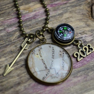 May include: A bronze necklace with a compass, a map charm, an arrow charm, and a 2023 charm. The map charm features a vintage map of Black Rock Desert, Nevada.