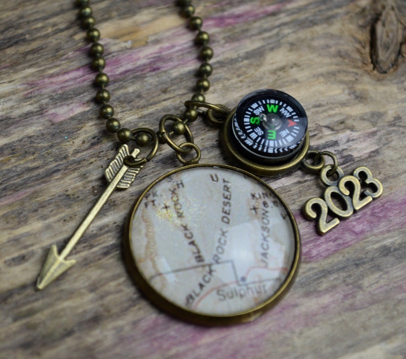 May include: A bronze necklace with a compass, a map charm, an arrow charm, and a 2023 charm. The map charm features a vintage map of Black Rock Desert, Nevada.