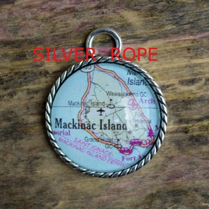 Op de afbeelding: Een zilveren bedel met een kaart van Mackinac Island, Michigan. De bedel is rond en heeft een touwachtige rand. De kaart toont de locatie van het Grand Hotel, de Wawashkimo Golf Course en Fort Mackinac.