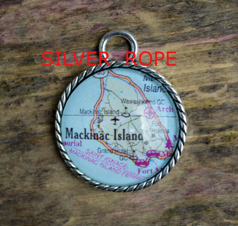 Op de afbeelding: Een zilveren bedel met een kaart van Mackinac Island, Michigan. De bedel is rond en heeft een touwachtige rand. De kaart toont de locatie van het Grand Hotel, de Wawashkimo Golf Course en Fort Mackinac.