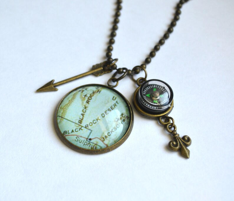 May include: A bronze necklace with a compass, arrow, and a glass cabochon featuring a map of Black Rock Desert. The map shows the words "Black Rock Desert", "Black Rock", "Sulphur", and "Jackson".