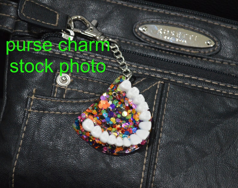 K&ouml;nnte beinhalten: Ein wei&szlig;er Zahnschmuck mit buntem Glitzer auf schwarzem Hintergrund. Der Anh&auml;nger ist an einer silbernen Kette und einem silbernen Verschluss befestigt.