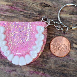 K&ouml;nnte beinhalten: Rosa glitzernder Schl&uuml;sselanh&auml;nger aus Harz mit wei&szlig;en Z&auml;hnen und einem silbernen Schl&uuml;sselring. Der Schl&uuml;sselanh&auml;nger liegt neben einem US-Cent zum Gr&ouml;&szlig;envergleich.