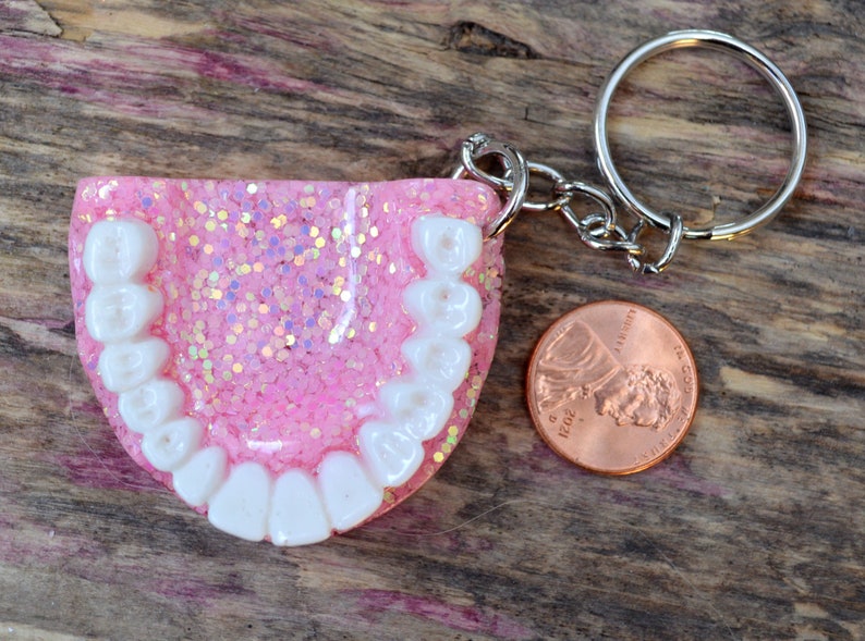 K&ouml;nnte beinhalten: Rosa glitzernder Schl&uuml;sselanh&auml;nger aus Harz mit wei&szlig;en Z&auml;hnen und einem silbernen Schl&uuml;sselring. Der Schl&uuml;sselanh&auml;nger liegt neben einem US-Cent zum Gr&ouml;&szlig;envergleich.