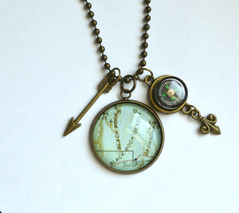 May include: A bronze necklace with a compass, arrow, and a glass pendant featuring a map of Black Rock Desert, Nevada. The map shows the words "Black Rock Desert", "Sulphur", and "Jackson".