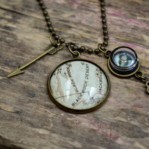 May include: A necklace with a compass, a key, and an arrow charm. The compass has a black face with a green needle. The key is a simple design with a clover-shaped head. The arrow charm is made of metal and has a brown finish. The necklace is made of a chain with a brown finish.