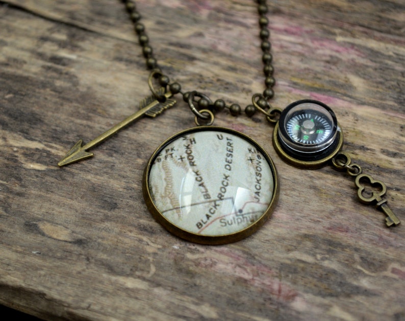 May include: A necklace with a compass, a key, and an arrow charm. The compass has a black face with a green needle. The key is a simple design with a clover-shaped head. The arrow charm is made of metal and has a brown finish. The necklace is made of a chain with a brown finish.