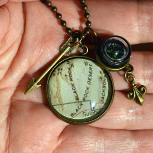 May include: A necklace with a compass, arrow, and a round pendant with a map of Black Rock Desert, Nevada. The pendant is made of clear resin with a white map inside. The necklace is made of a brass chain.