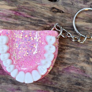 K&ouml;nnte beinhalten: Rosa glitzernder Schl&uuml;sselanh&auml;nger aus Harz in Form eines Gebisses mit wei&szlig;en Z&auml;hnen.