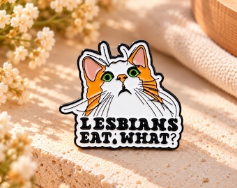 May include: A white and orange cat enamel pin with green eyes and a confused expression. The text below the cat reads LESBIANS EAT WHAT? in bold black letters. The pin features a black metal outline and a white background.