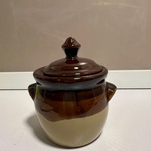 Può includere: Un barattolo in ceramica con coperchio, caratterizzato da un design a due tonalità. La parte superiore e il coperchio sono di un marrone scuro lucido, mentre il corpo inferiore è di un beige opaco liscio. Il barattolo ha due piccole maniglie.