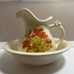 Puede incluir: Juego de jarra y cuenco de cerámica color crema. La jarra presenta un asa y un pico, con un diseño pintado de uvas verdes, nueces marrones y hojas naranjas. El cuenco es redondo y se encuentra debajo de la jarra.