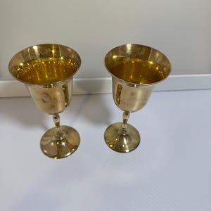 Puede incluir: Dos copas doradas vintage con un borde ancho y acampanado y un tallo delgado. Las copas tienen una superficie brillante y reflectante y se asientan sobre una superficie blanca. Las copas probablemente son de metal y son adecuadas para servir bebidas.