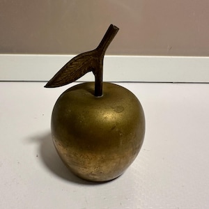 May include: A brass apple sculpture with a leaf. The apple is a golden color, and the leaf and stem are a darker bronze. The sculpture is a decorative item, likely for a table or shelf.