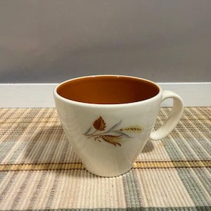May include: A cream-colored ceramic mug with a wide, tapered body and a handle on the right side. The interior of the mug is a solid burnt orange color. A botanical illustration of brown leaves and wheat stalks is printed on the exterior surface.