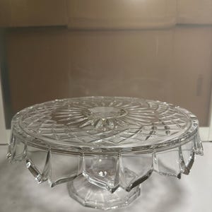 May include: A clear glass cake stand with a decorative, diamond-cut pattern. The stand has a round top with a scalloped edge and a pedestal base. The glass reflects light, creating a sparkling effect. Ideal for displaying cakes or desserts.